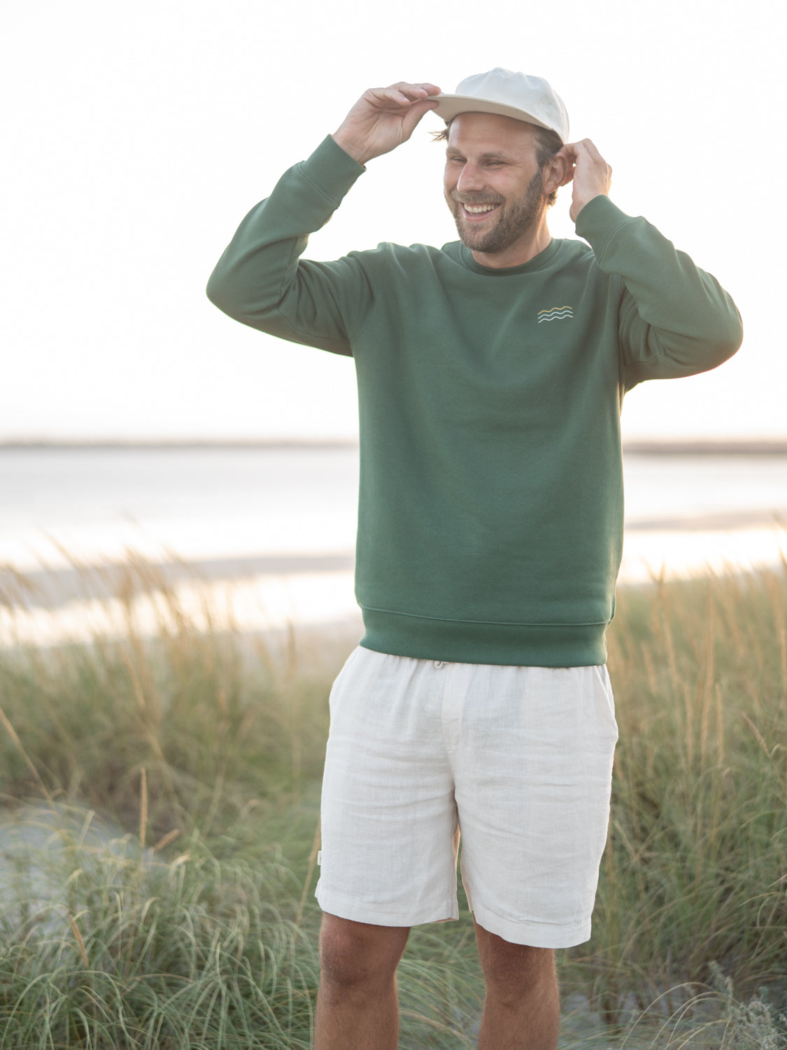 Nachhaltiger Fair Fashion Unisex Pullover von FUXBAU in grün mit einer Wellen Stickerei aus Biobaumwolle getragen von Philipp am Strand in Portugal.