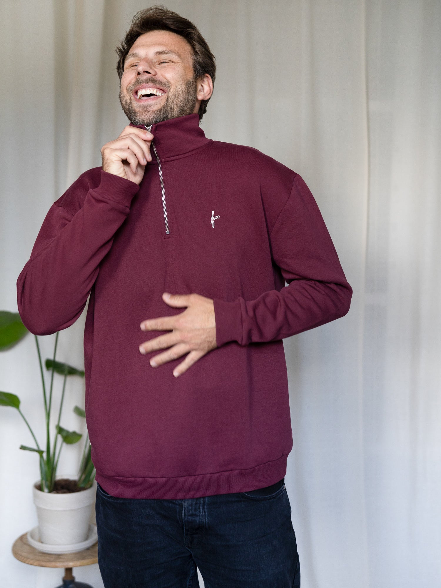Mann zieht den Reißverschluss des bordeauxfarbenen FUXBAU Half-Zip Sweaters hoch – nachhaltiger Pullover aus Biobaumwolle.