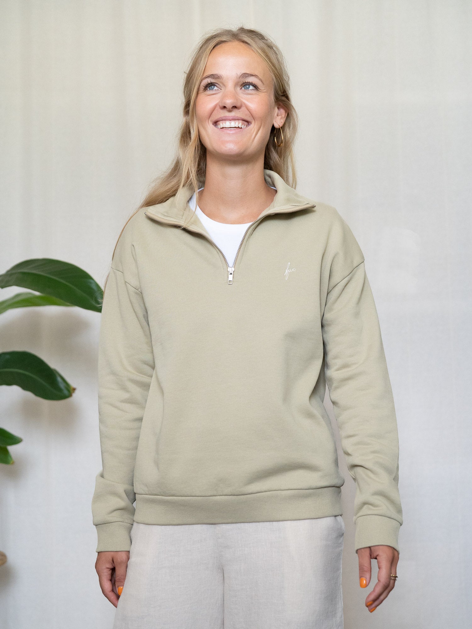 Person trägt salbeifarbenen Half-Zip-Sweater aus Biobaumwolle, steht entspannt mit leichtem Lächeln
