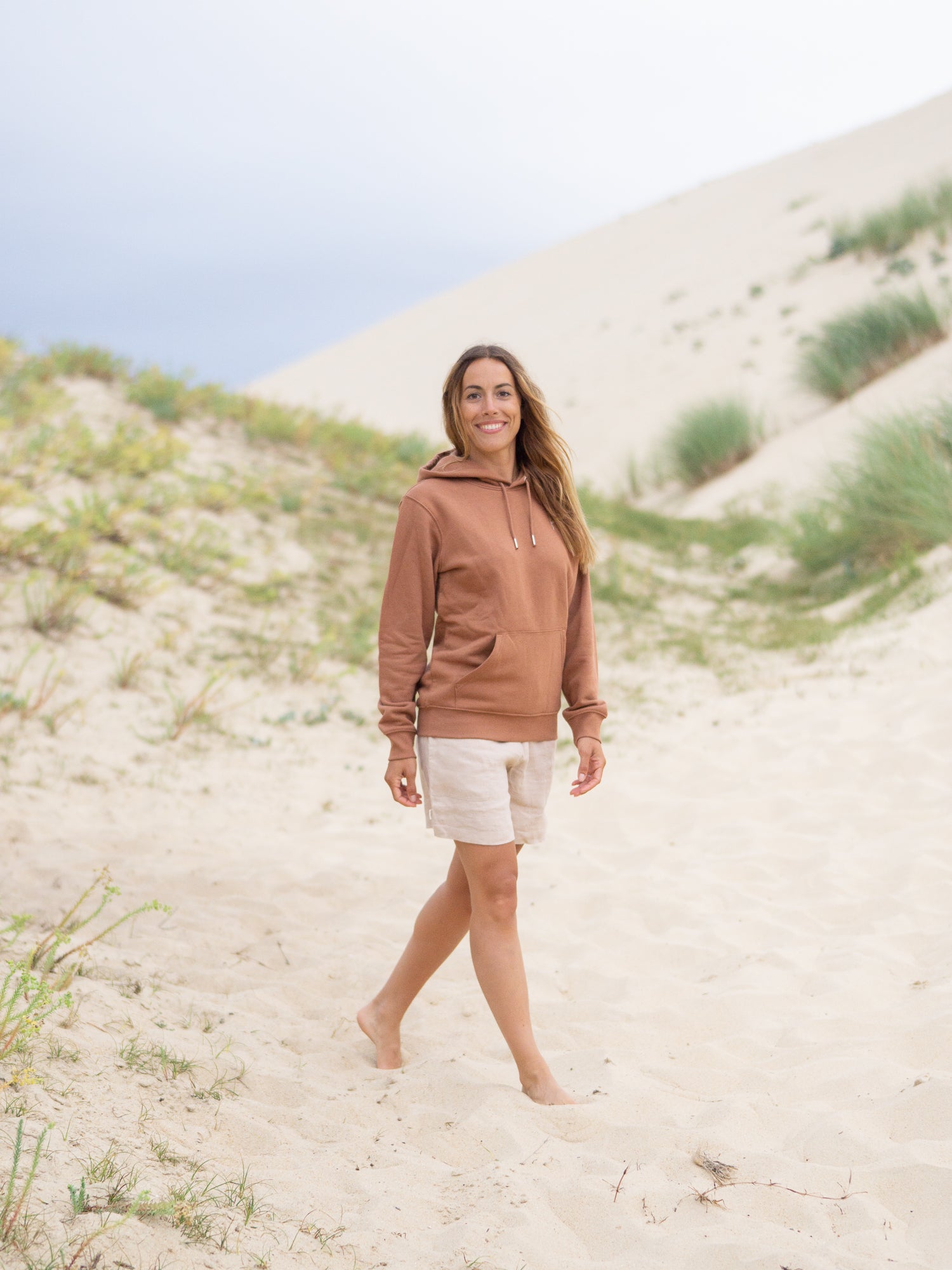 Unsere Freundin Rachel trägt unseren unisex Fair Fashion Hoodie in karamell am Strand aus 100% GOTS zertifizierter Biobaumwolle und made in Portugal