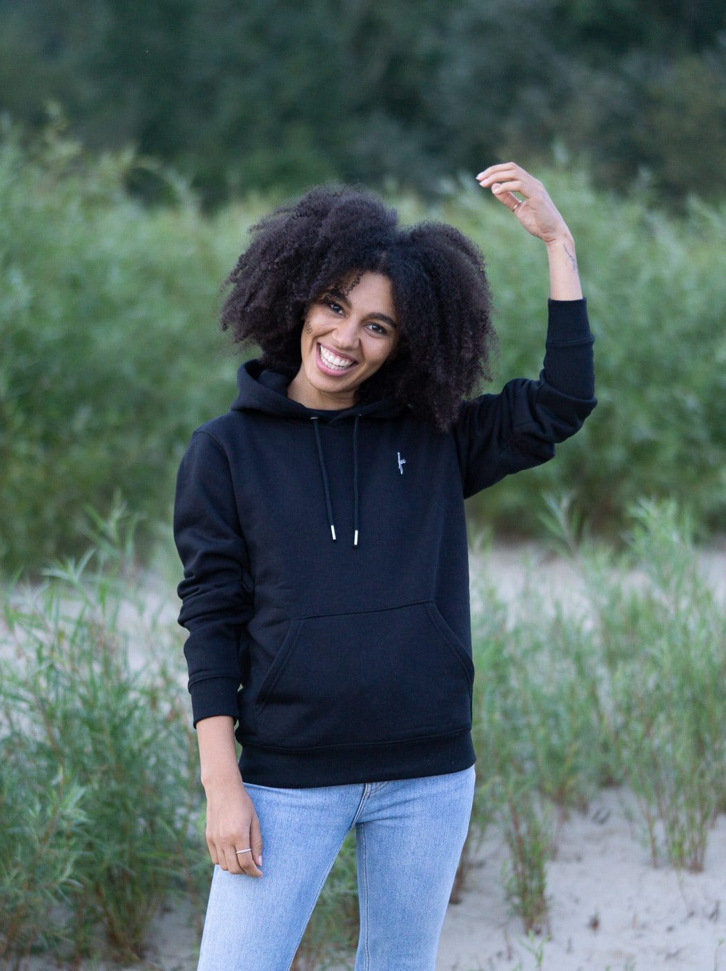 Junge, schwarzhaarige Frau trägt einen fairenund nachhaltigen Hoodie von FUXBAU in schwarz in Hamburg am Strand.